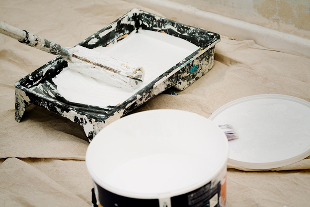 A paint roller and tray with white paint, alongside a bucket and lid, on a protective drop cloth.